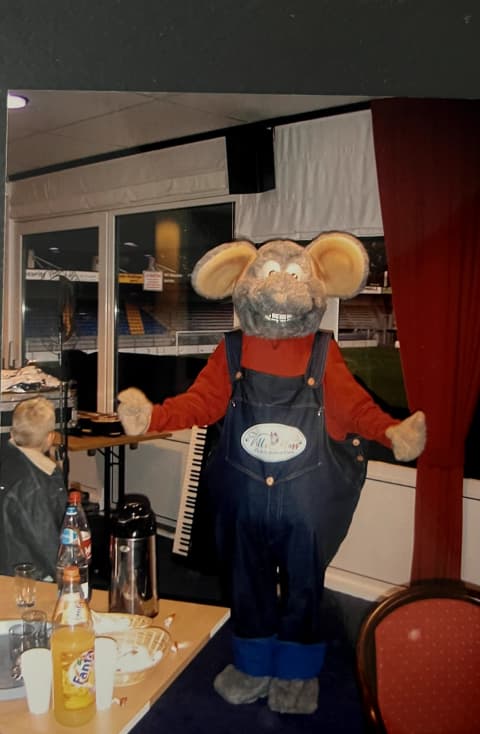 Arjo the Rat, de mascotte van Villa Happ, in tuinbroek met het merklogo.